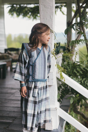 Young girl in a plaid kimono standing on a wooden deck with greenery in the background