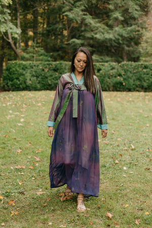 Woman wearing a purple shot silk han bok dress and jacket.  Jacket is of teal and green shot silk and tied in the front.  Woman is walking in a lawn with shrubs in the background.