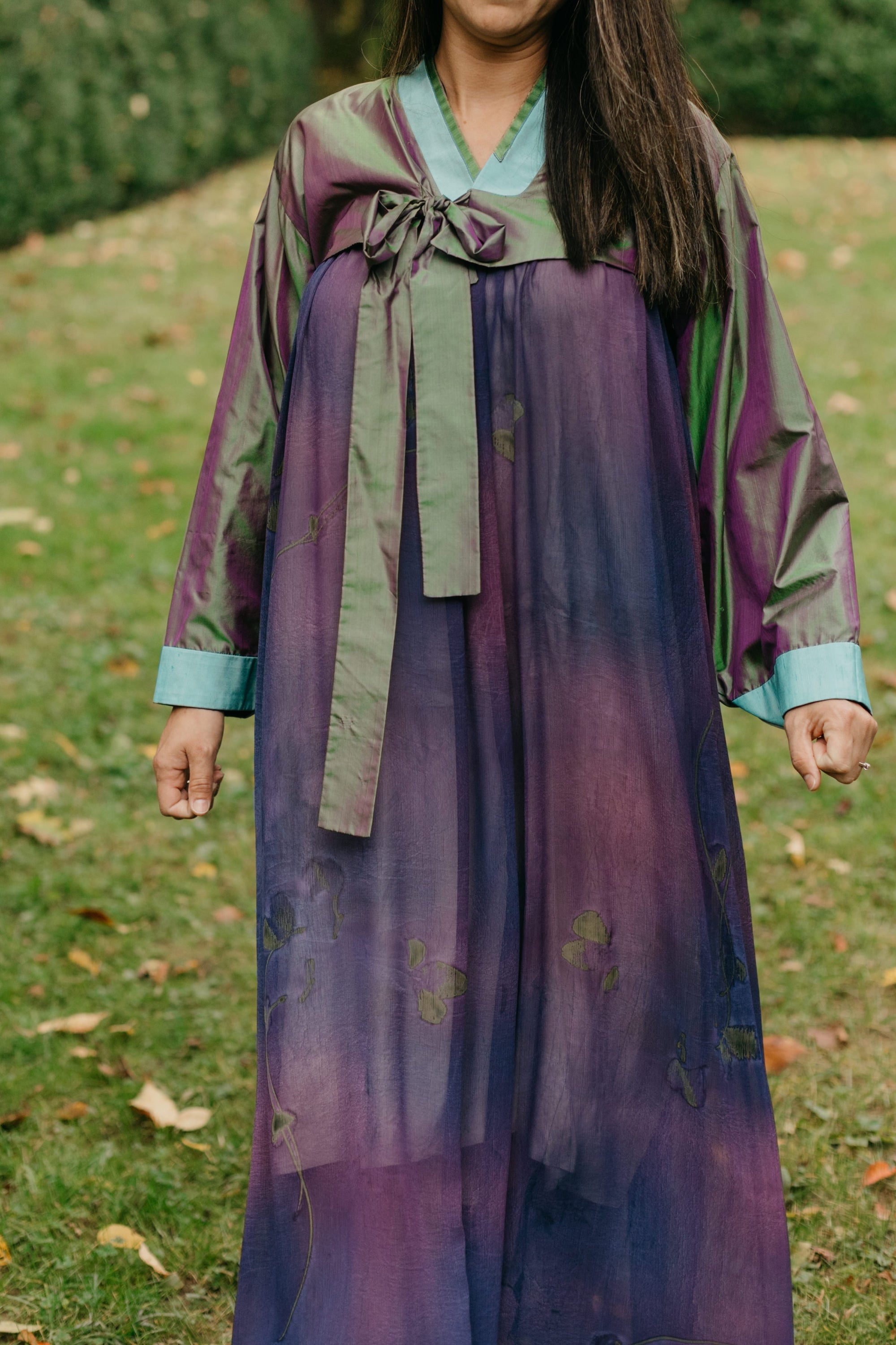 Woman wearing a purple shot silk han bok dress and jacket. Jacket is of teal and green shot silk and tied in the front. She is outside in a lawn.  Photo is close up of the front of the hanbok..