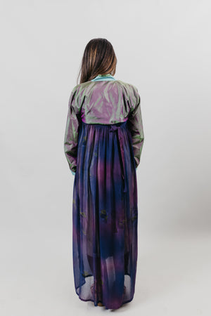 Back View of a woman wearing a purple shot silk han bok dress and jacket. Jacket is of teal and green shot silk and tied in the front. She is standing in front of a white backdrop.