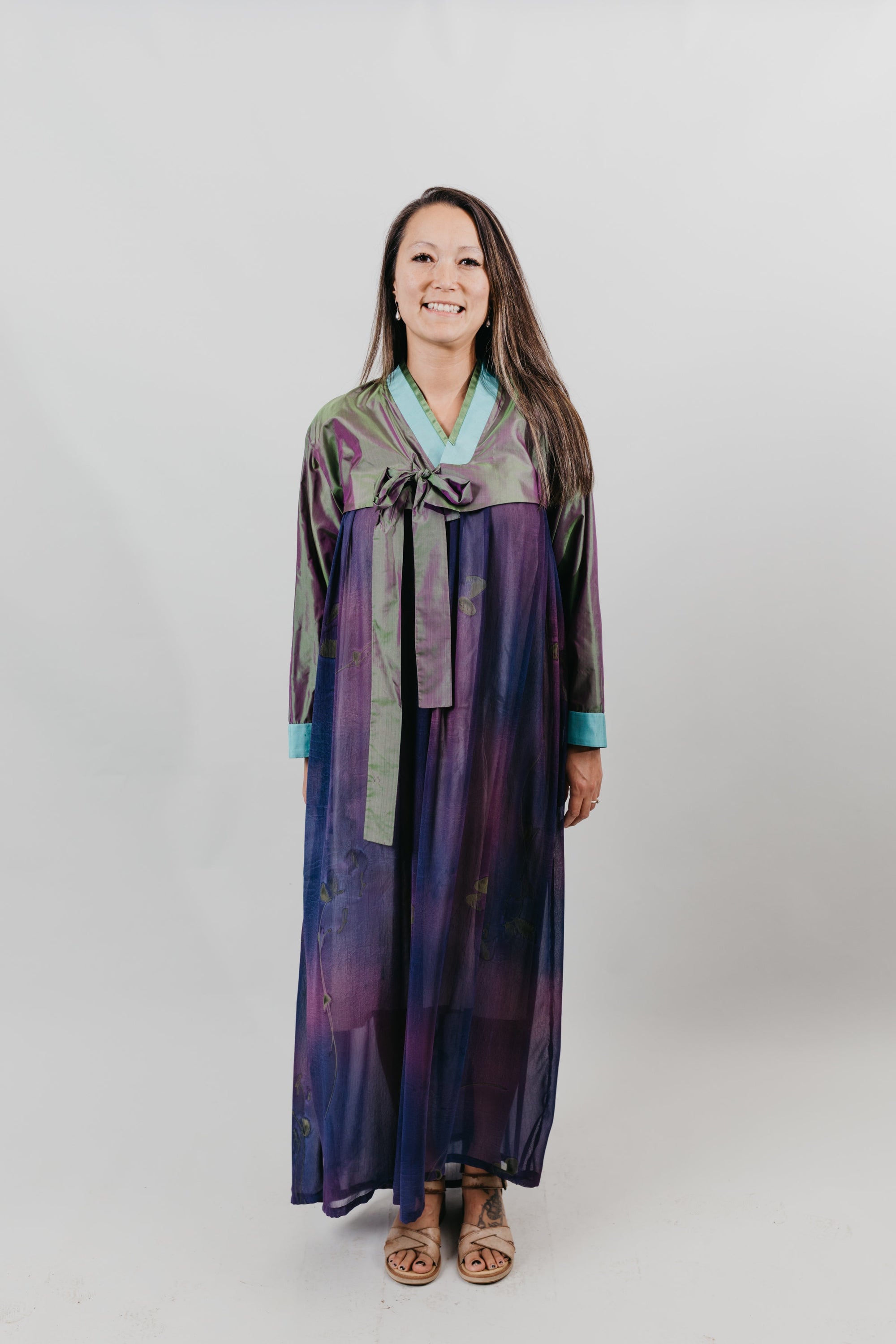 Woman wearing a purple shot silk han bok dress and jacket. Jacket is of teal and green shot silk and tied in the front. She is standing in front of a white backdrop.