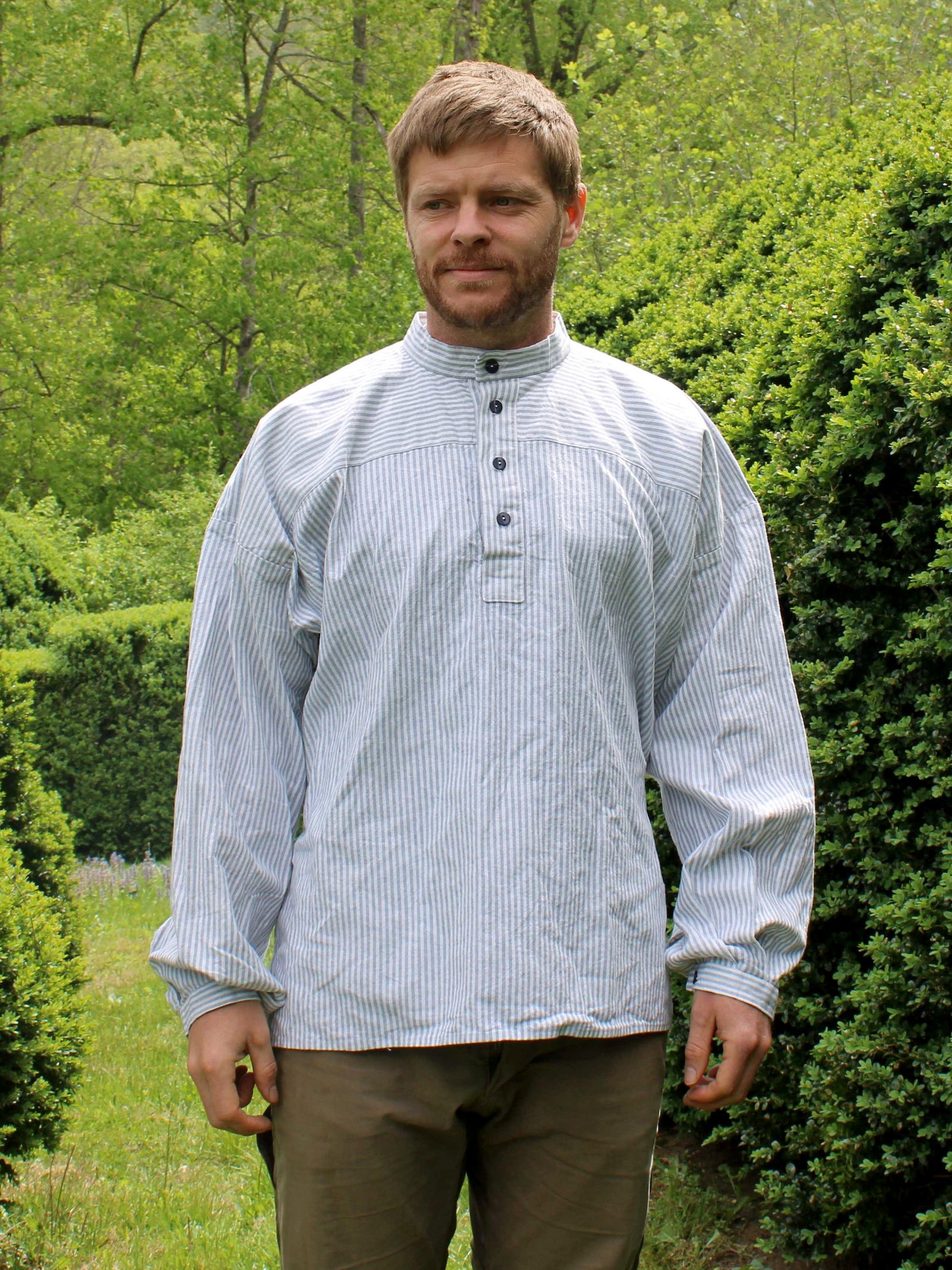 Man wearing a blue and white striped busserull shirt outside by green shrubbery
