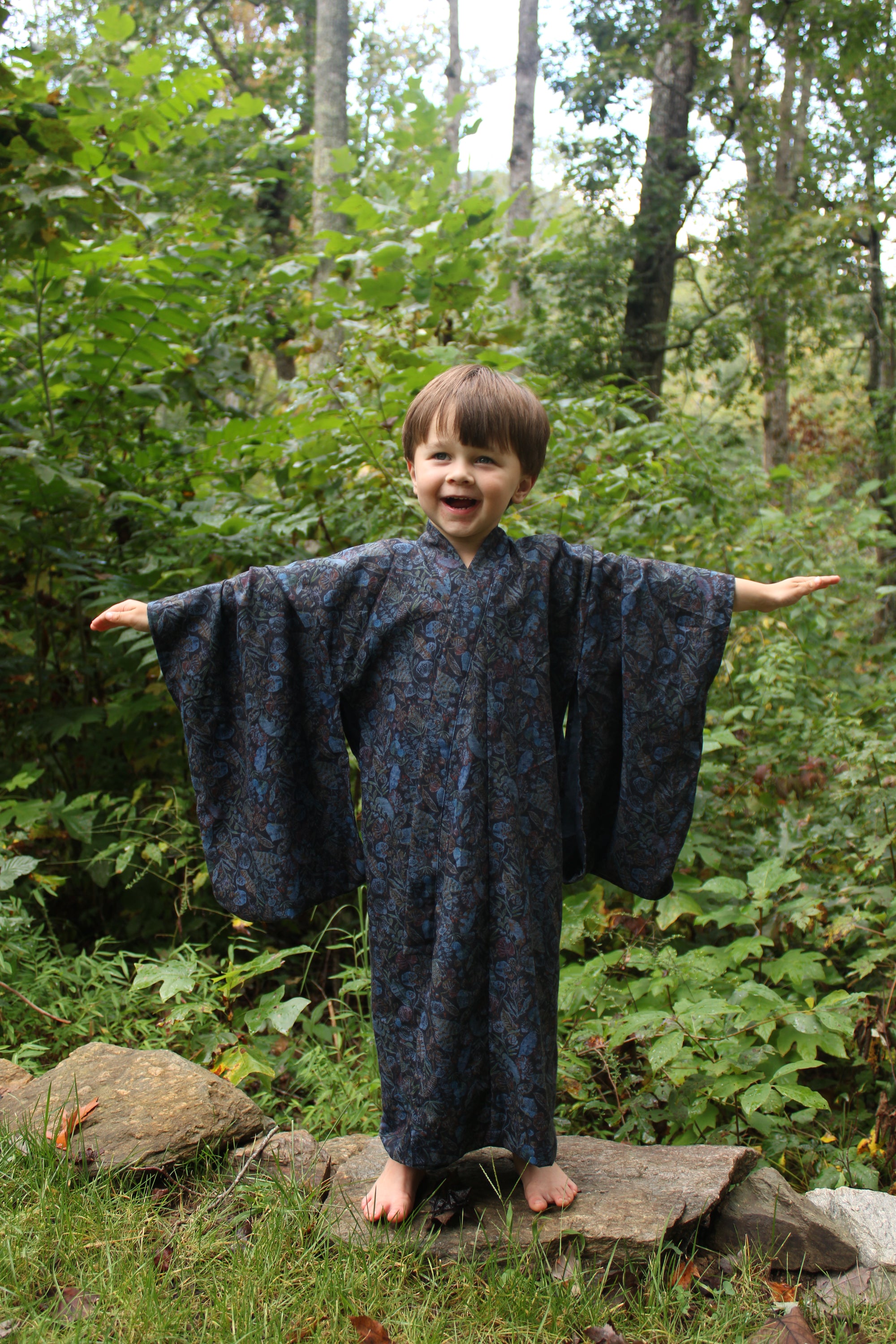 Boy standing outdoors in a forest in  a dark blue kimono with trees and greenery in the background