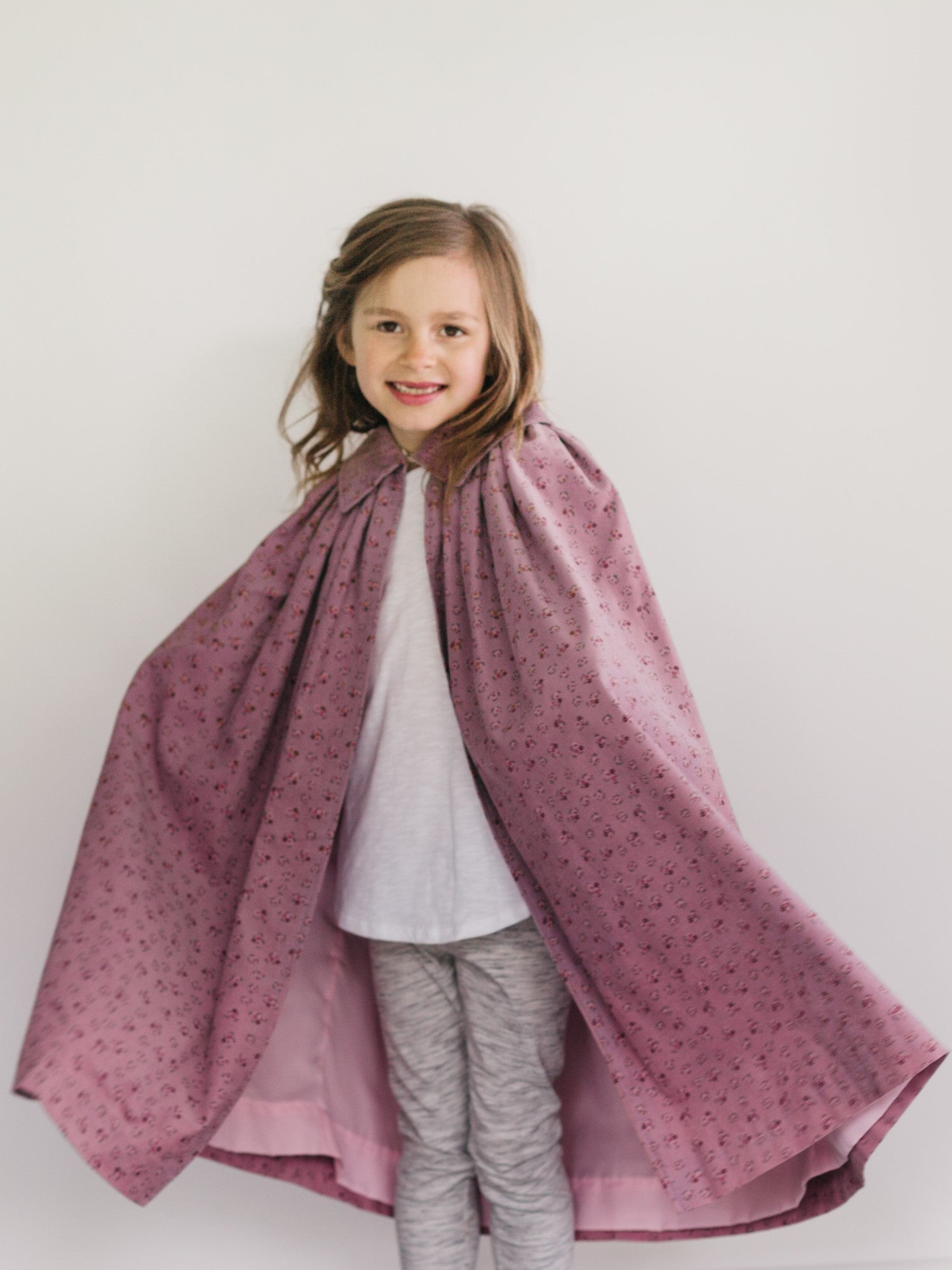 Young girl wearing a pink printed kinsale cloak or cape with a collar.  Grey pants and white shirt under the cape.
