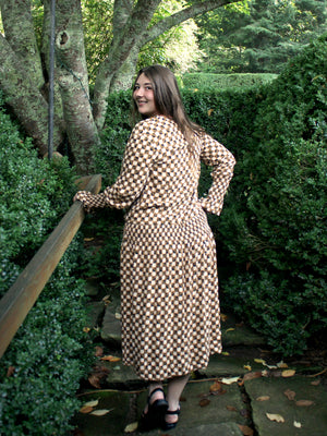 Woman by bushes wearing a long sleeved patterned tea frock with her back to the camera