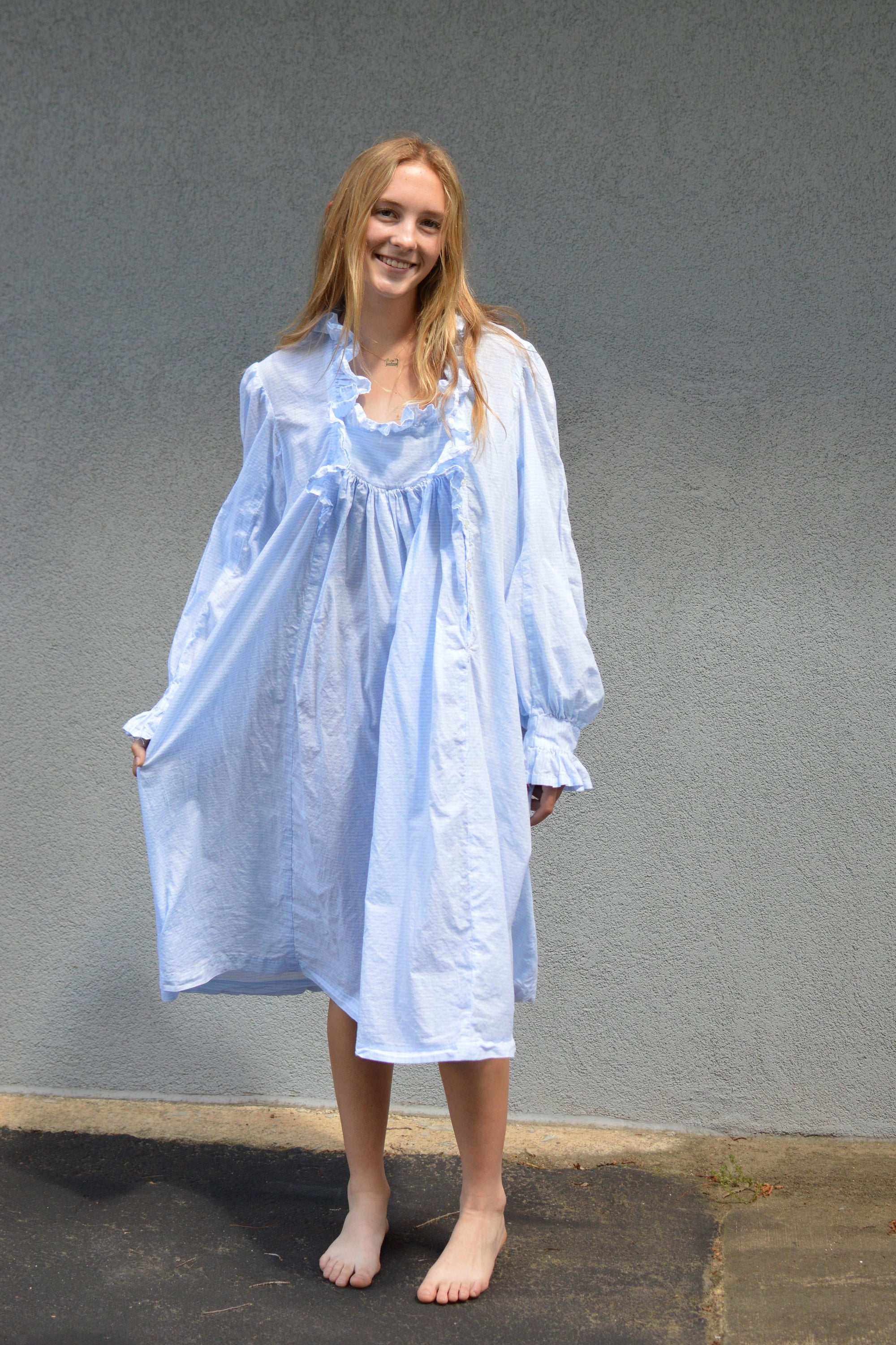 Young woman standing in front of a grey wall in a light blue ruffled nightgown.