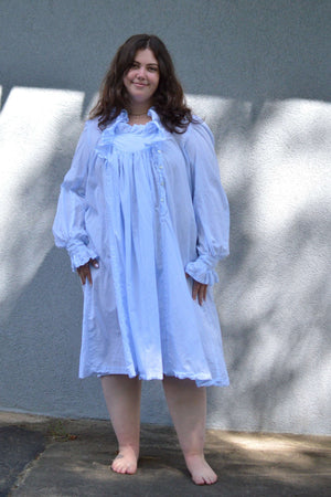 Young woman standing in front of a grey wall in a light blue ruffled nightgown.