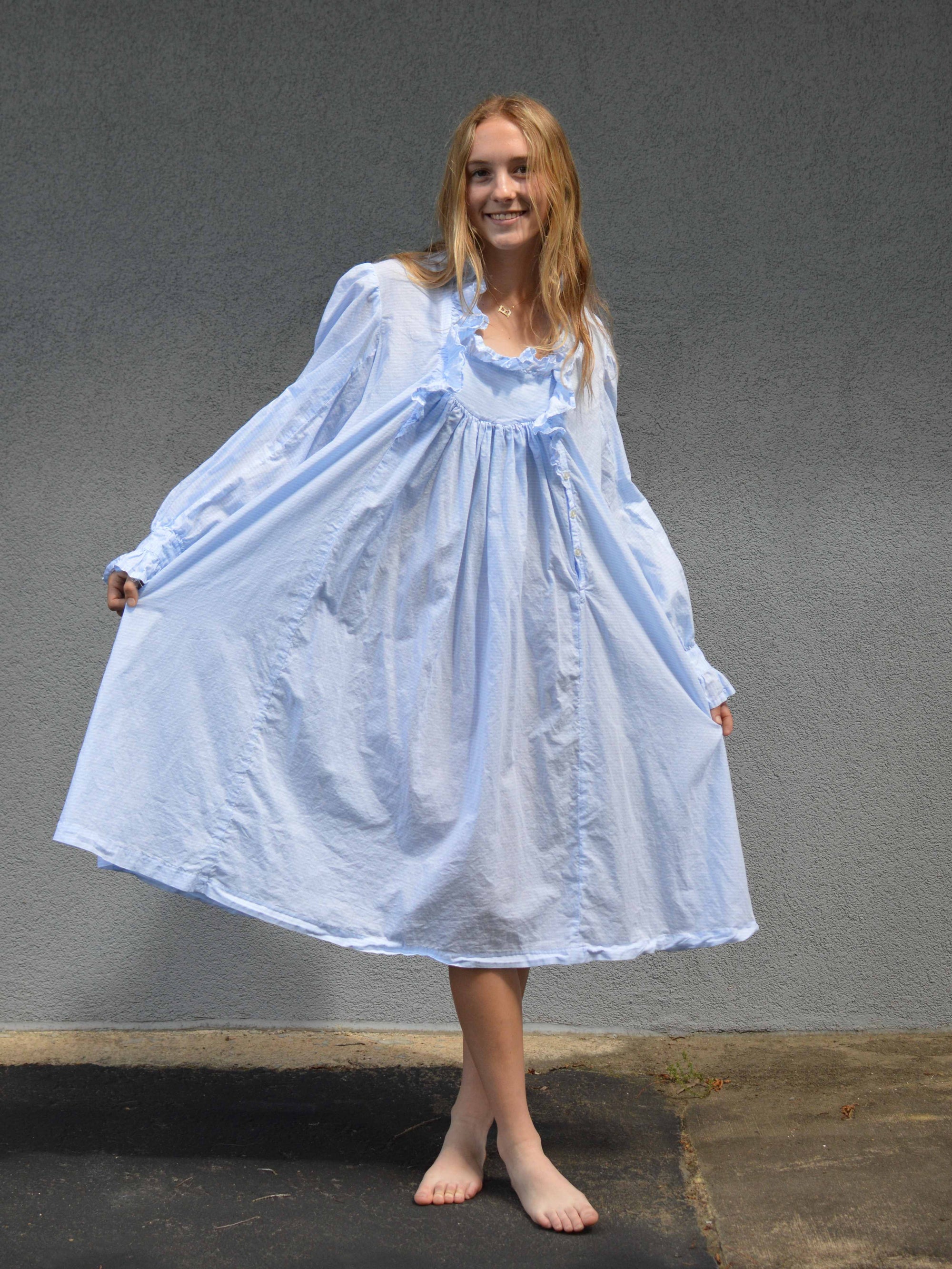 Young woman standing in front of a grey wall in a light blue ruffled nightgown.