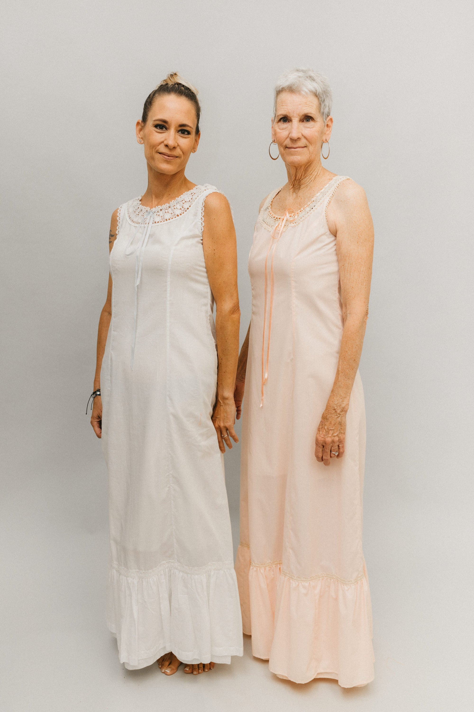 Two women wearing floor length princess slips with lace at the necklines.  Slip on right is light peach, slip on left is white.