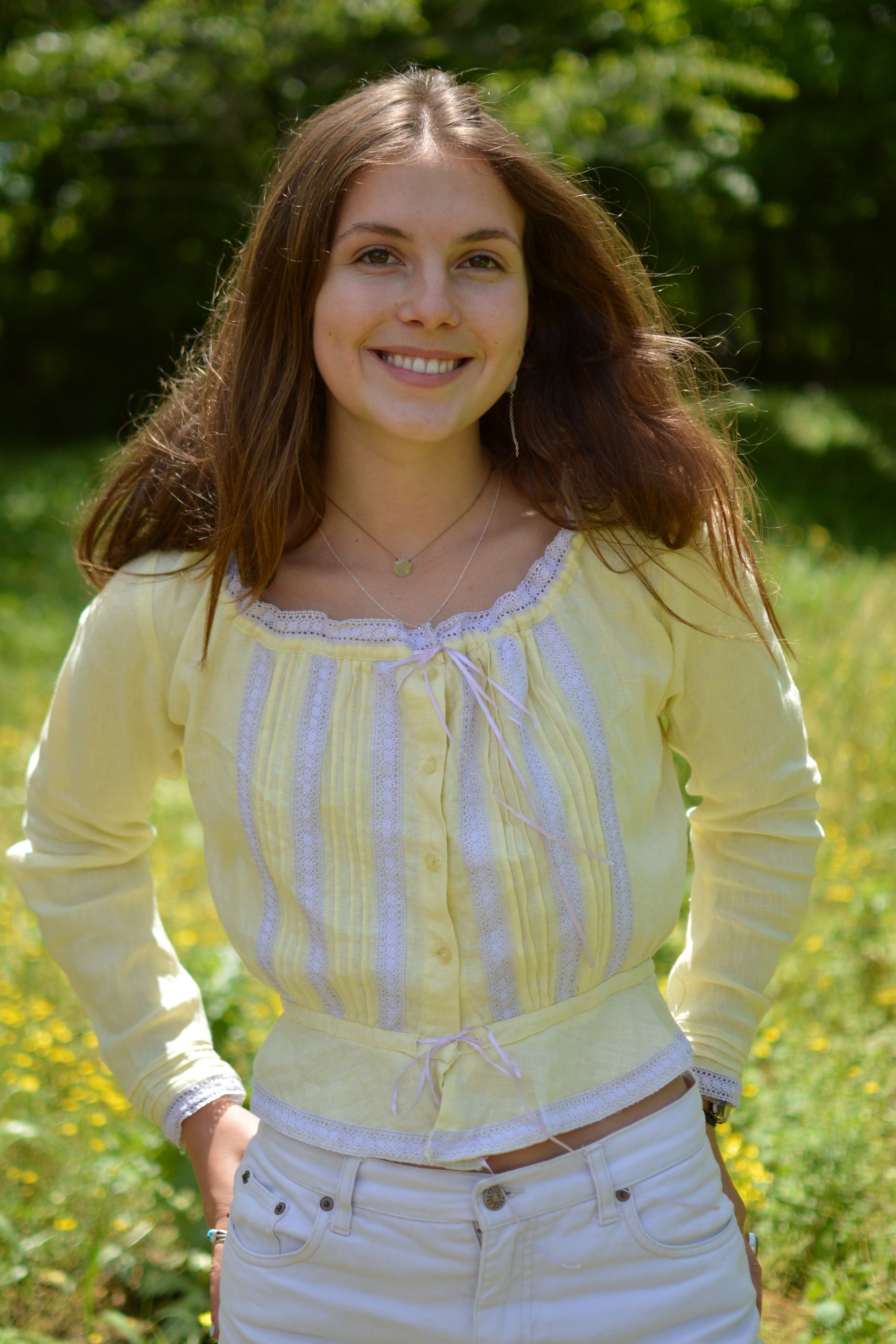 young white woman wearing a pastel long sleeved camisole with pastel purple lace. hands in the back of her pockets and smiling. With greenery in the background.