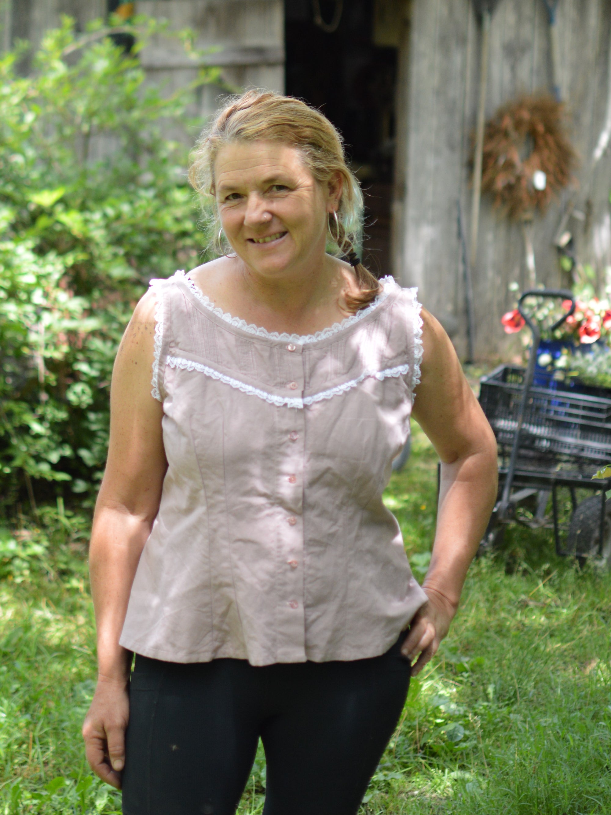 smiling middle aged white blonde woman standing with left hand on her hip wearing sleeveless camisole in pale pink with white ruffle lace and black athletic leggings with greenery in the background