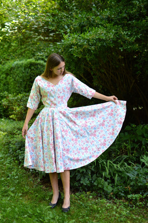 Young woman standing in front of trees and shrubs wearing a white and pink floral print dress with a v-neck.  She is holding one edge of the dress skirt up. 