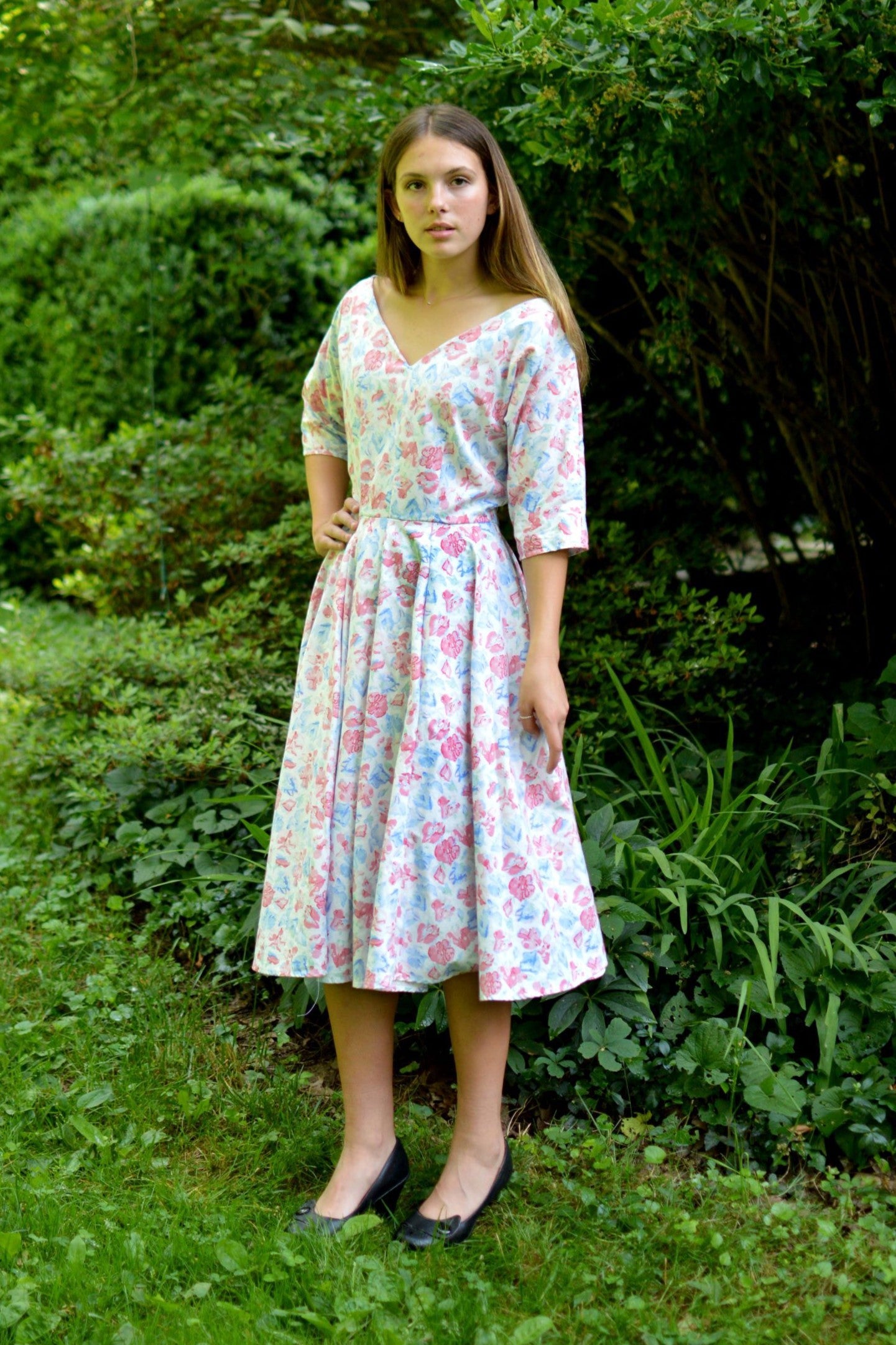 Young woman standing in front of trees and shrubs wearing a white and pink floral print dress with a v-neck.