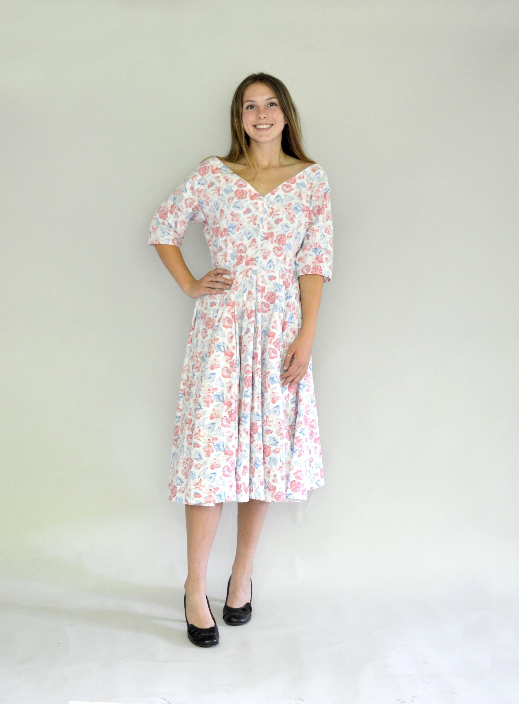 Young woman smiling wearing a below-knee length white and pink floral print dress with a v-neck and 3/4 length sleeves and black shoes.  She is in front of a white background.