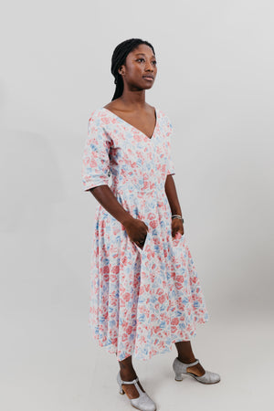 Young woman wearing a below-knee length white and pink floral print dress with a v-neck and 3/4 length sleeves and black shoes.  She is in front of a white background and her hands are in the skirt pockets.