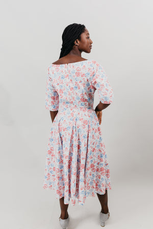 Young woman with her back to viewer is wearing a below-knee length white and pink floral print dress with a v-neck and 3/4 length sleeves and black shoes.  She is in front of a white background.