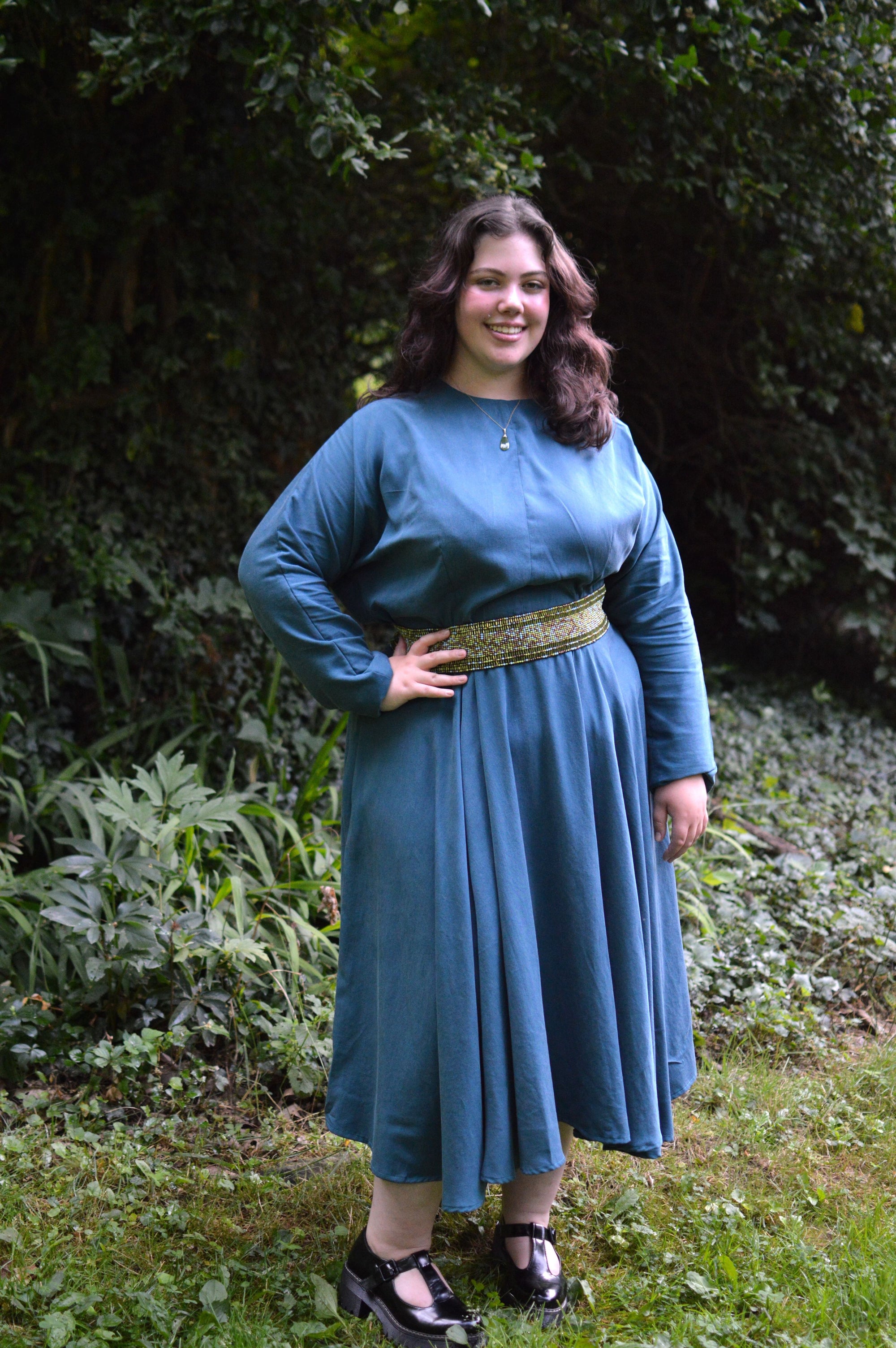 Young woman wearing a blue long sleeve, jewel neck, calf-length dress outside in front of shrubbery.  She has on a green a white beaded belt and black shoes. Her right hand is on her hip.