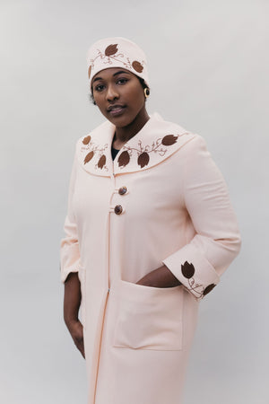Black woman wearing a pink wool coat and cloche with brown leaf applique and embroidery at the cuffs, collar, and hat.