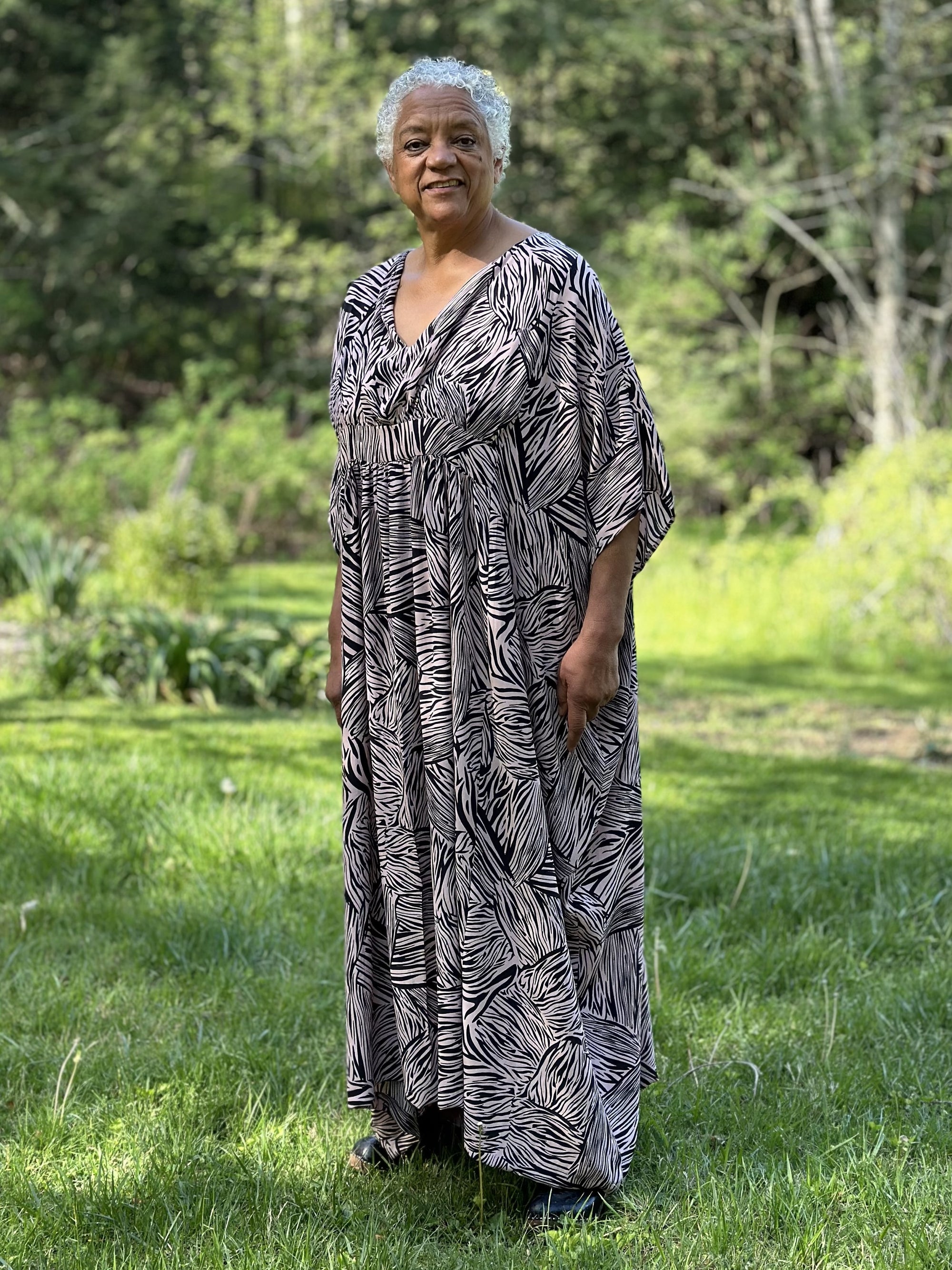 Standing older African American woman wearing Kaftan in a black and pink stripe like print with greenery in the background, with her hand on her hip and the other hand holding out the dress.