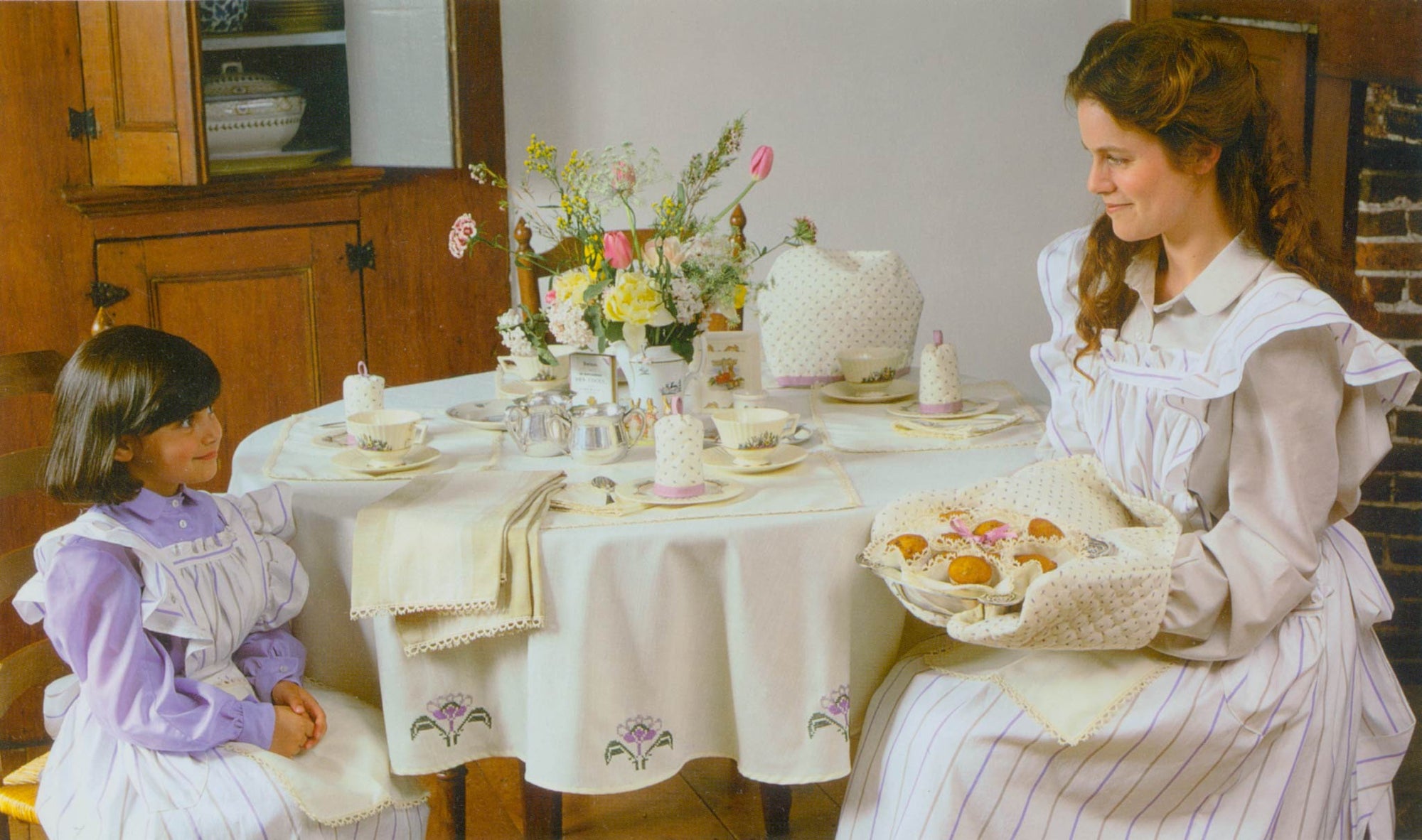 A young girl and woman sitting at a table with the tea . egg and biscuit cozy as well as the table cloth, napkins and placemats on the table. Both people are wearing the apron in a white and blue striped fabric looking at each other.