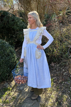 A woman standing outside with greenery in the background. She is wearing the  blue linen apron with beige ruffled straps , pockets and waistband. She has her left hand on her hip and a basket of eggs being held with her right hand