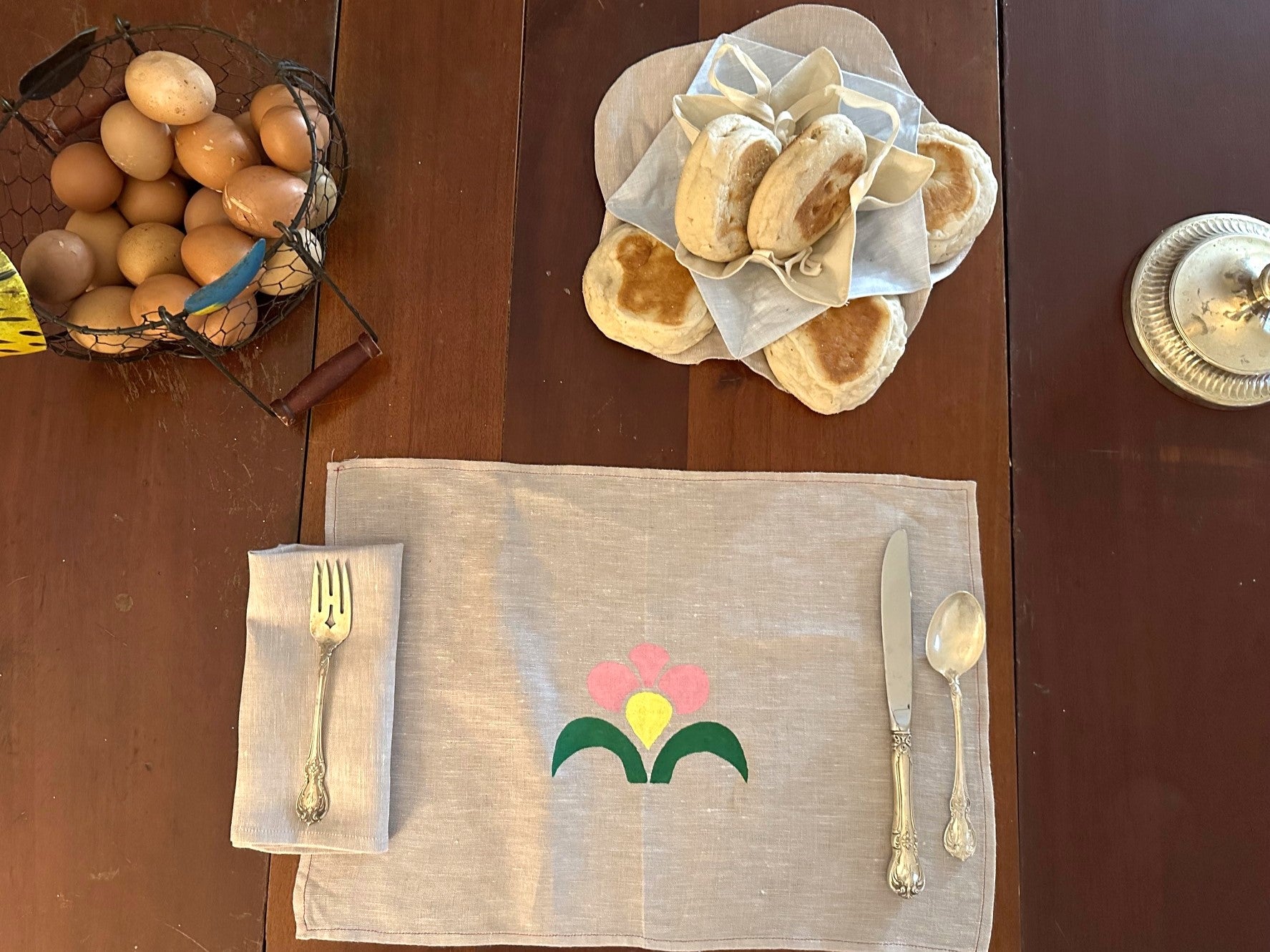 On a wooden table is a basket of eggs on the left top corner, biscuits in a biscuit cozy on the center top, a silver candle stick on the right top corner. On the center bottom there is a napkin with a silver fork on top, a placemat with a painted flower in pink yellow and green with a knife and spoon on top.