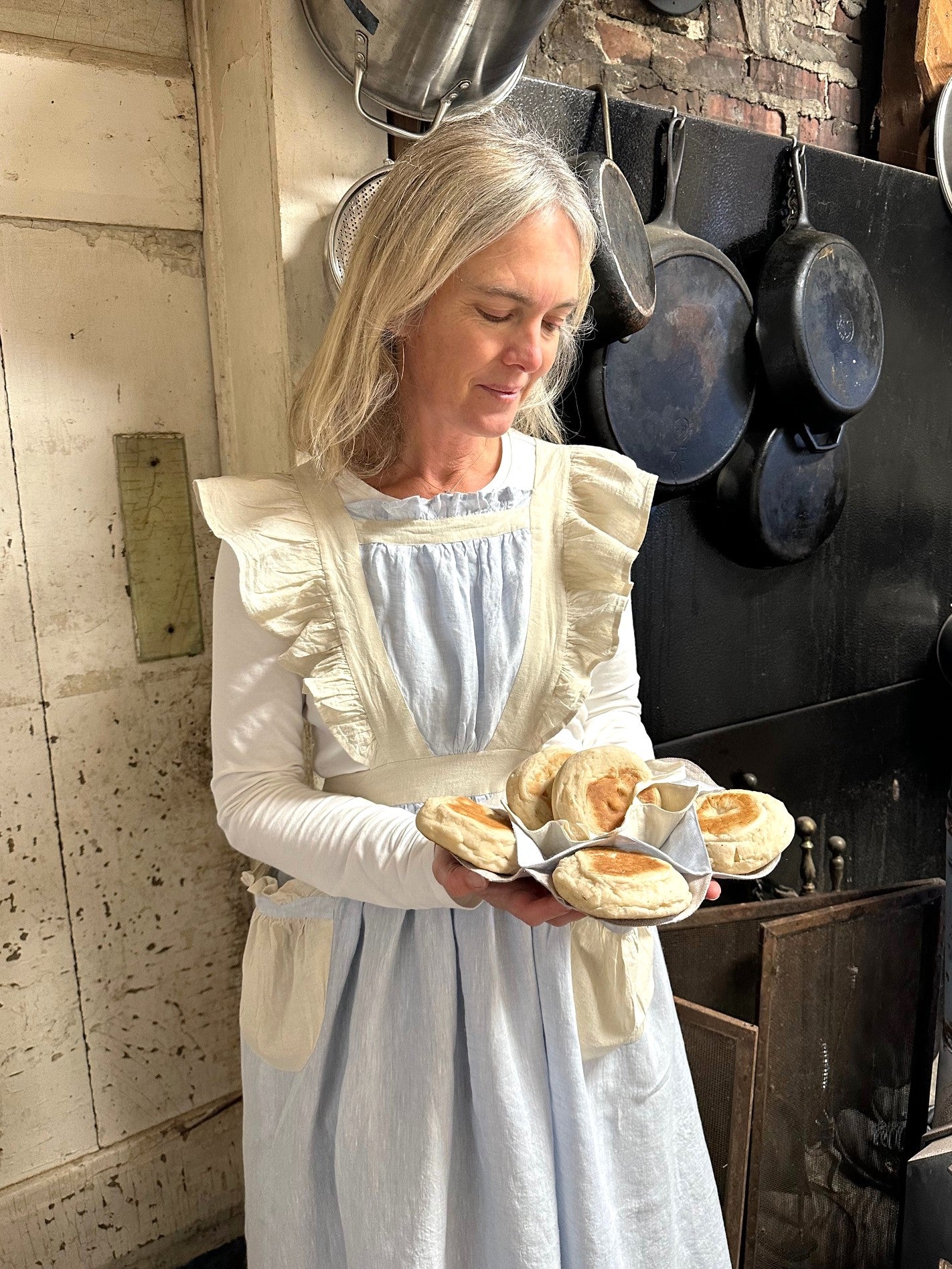 A woman wearing the blue linen apron with beige ruffled straps , pockets and waistband. She is holding biscuits in the biscuit cozy looking down at them. She is inside a kitchen with pots and pans in the background.