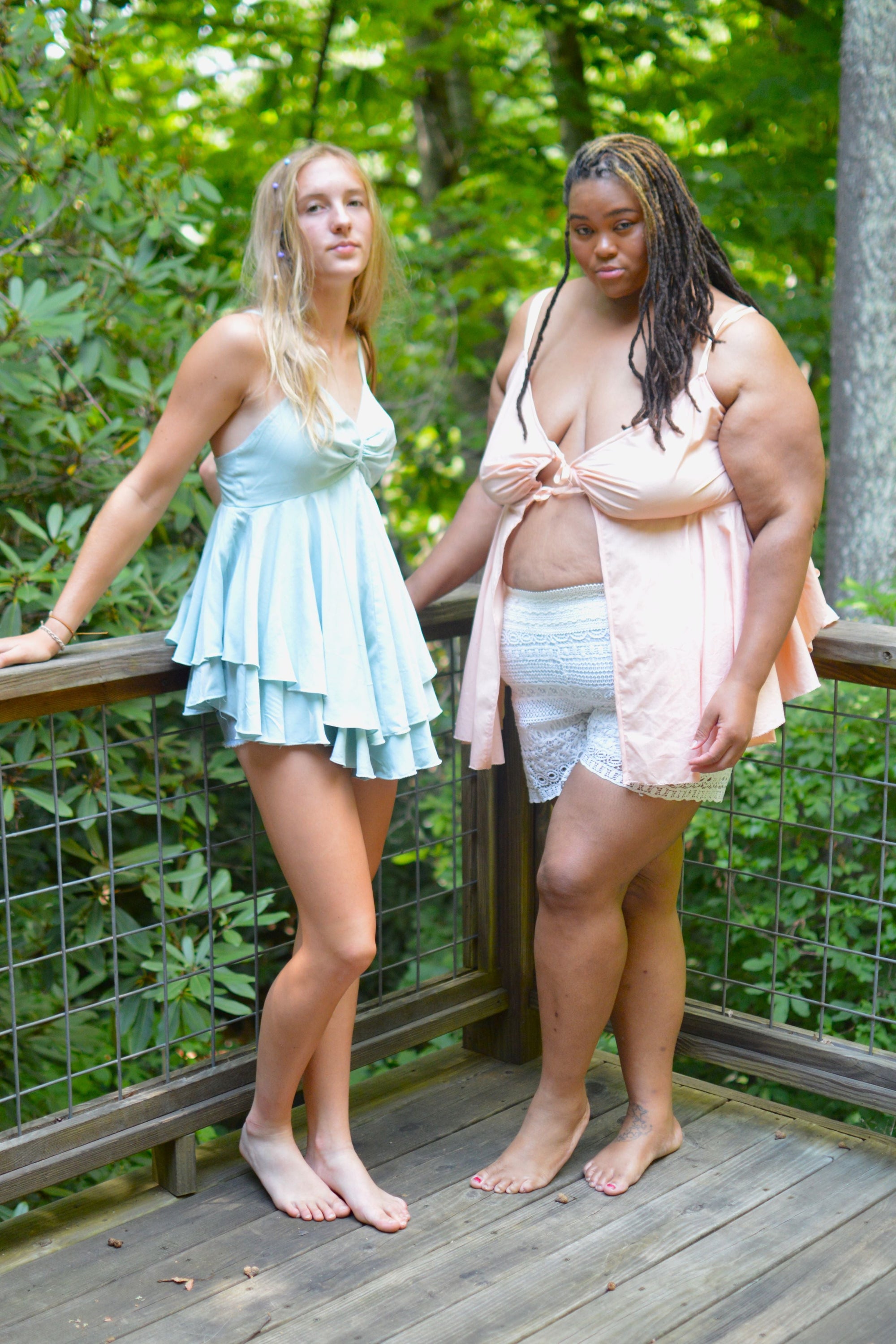 Two woman standing on a wooden porch with forest background.  One is wearing a light blue lingerie dress and the other is wearing a peach colored lingerie top that is open in center front.