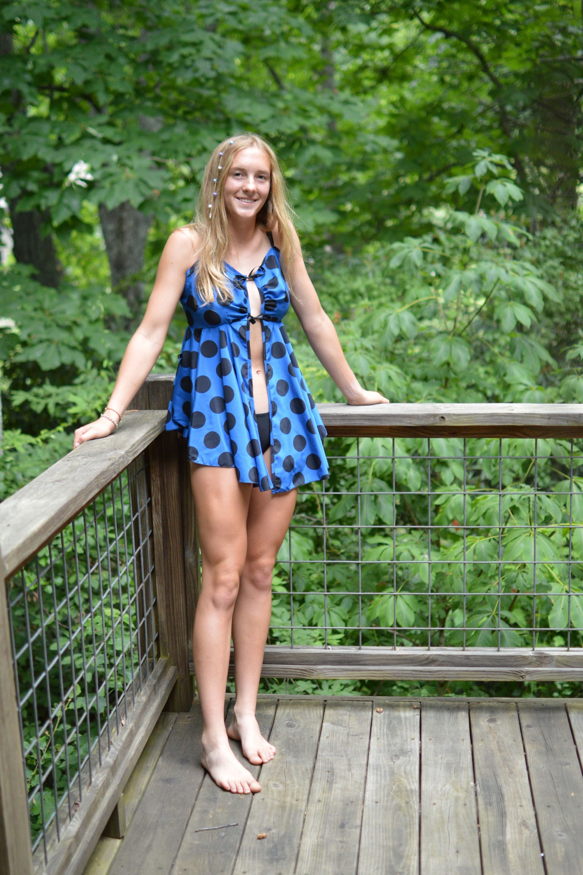 Woman standing on a wooden porch in the woods wearing a blue and black polka dot lingerie dress that is open in the center front and tied with black bows.