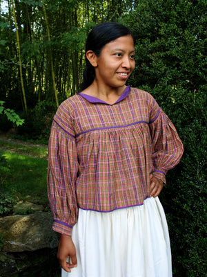 A woman wearing a purple and orange plaid long-sleeve blouse with a white skirt, standing outdoors in front of a green forest background.