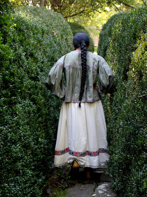 A woman viewed from behind wearing a long-sleeved vintage-style dress with a dark green jacket and an off-white skirt, walking down a stone path between tall green hedges.