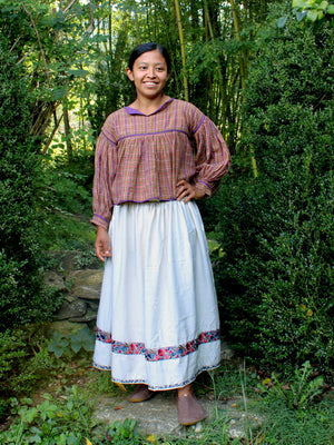 A woman standing outdoors in a lush green garden, wearing a traditional long-sleeve plaid blouse in purple and brown tones with a long white skirt featuring a colorful floral border.