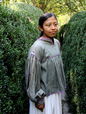 A woman standing outdoors between tall green hedges, wearing a loose gray long-sleeve blouse with purple trim and a white skirt.