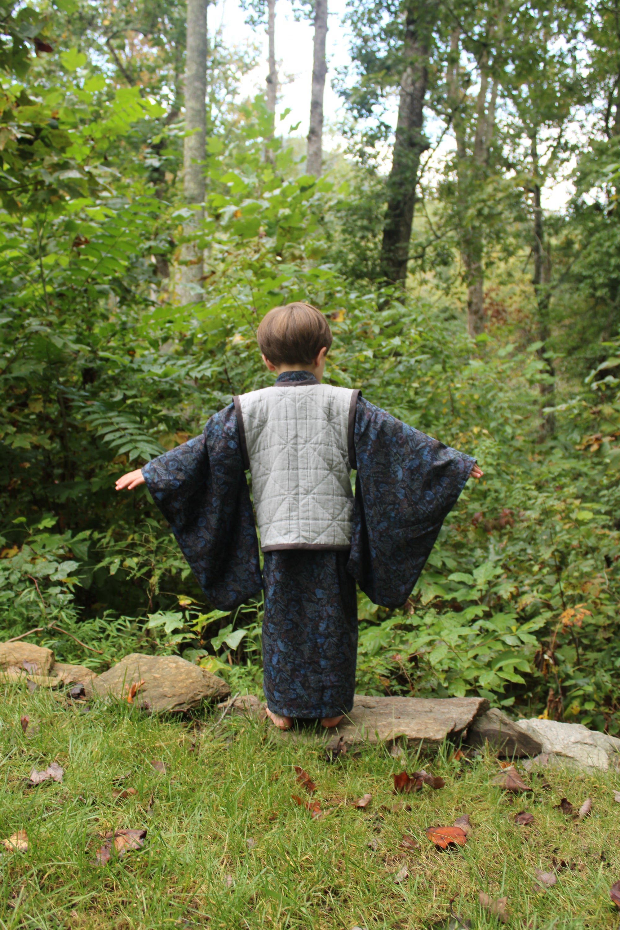 Boy with back to camera standing in a forest with trees and greenery in the background