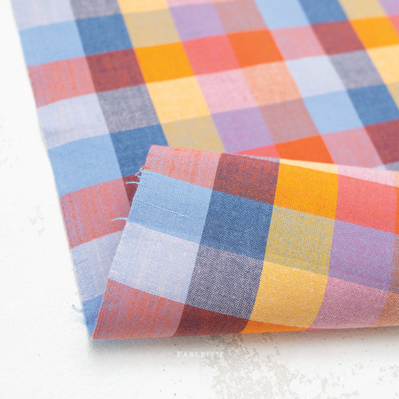 A colorful100% cotton fabric in a bold, woven check pattern in warm oranges, corals, yellows, soft pinks, and cool blues, on a white background.
