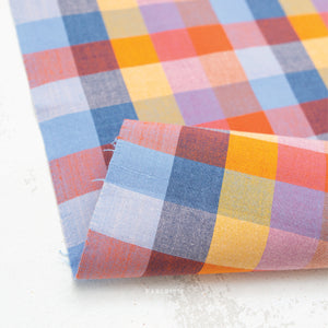 A colorful100% cotton fabric in a bold, woven check pattern in warm oranges, corals, yellows, soft pinks, and cool blues, on a white background.
