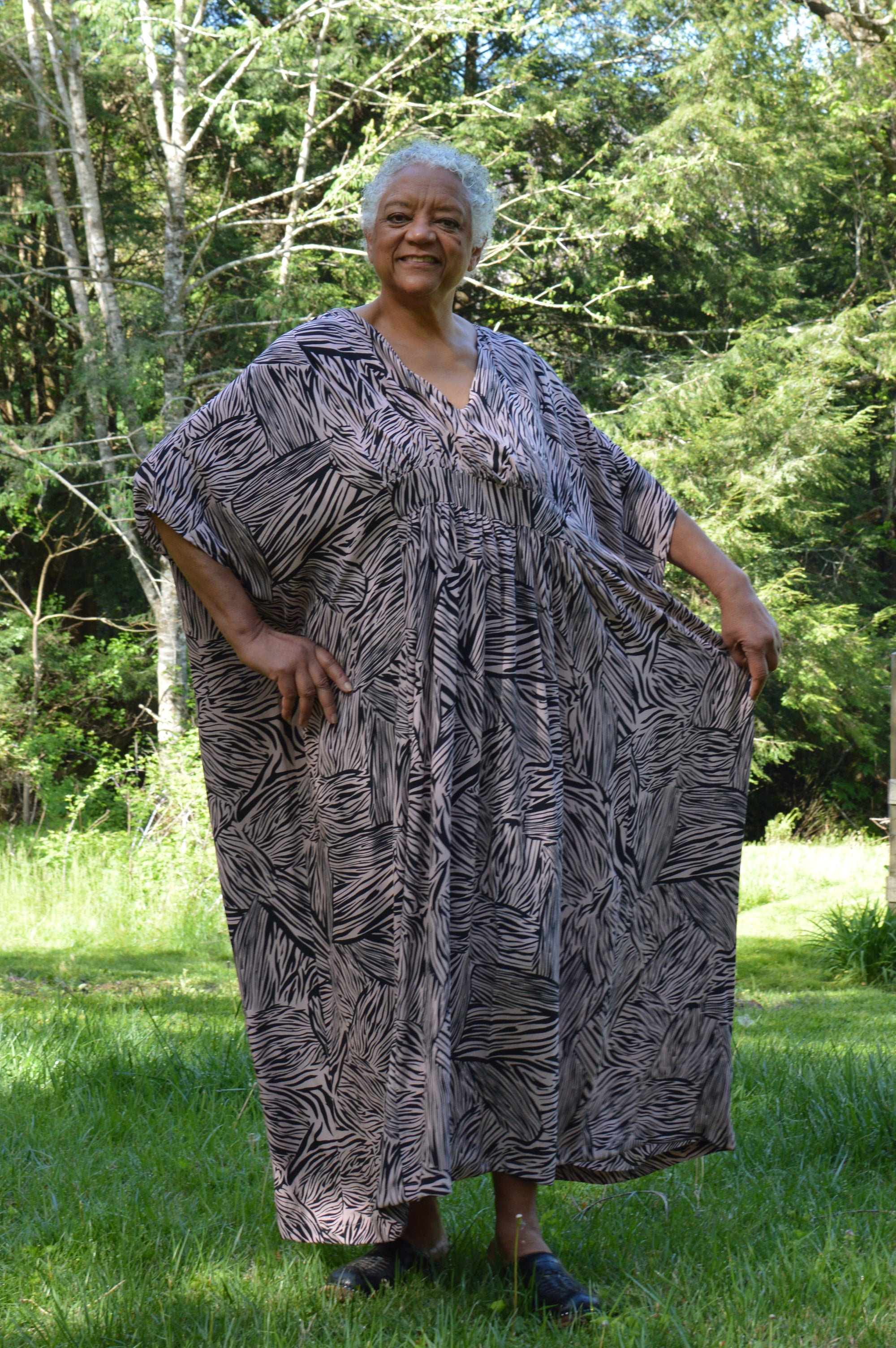 Standing older African American woman wearing Kaftan in a black and pink stripe like print with greenery in the background, with her hand on her hip and the other hand holding out the dress.