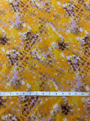 Yellow fabric with watercolor splashes in brown, purple, and white, measured by a ruler at the bottom.