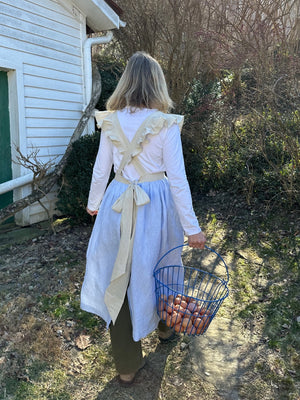 Outside in greenery the back of a woman in a white long sleeved shirt and green pants wearing the light blue linen apron with beige ruffled straps , pockets and waistband holding a basket of eggs in her right hand.