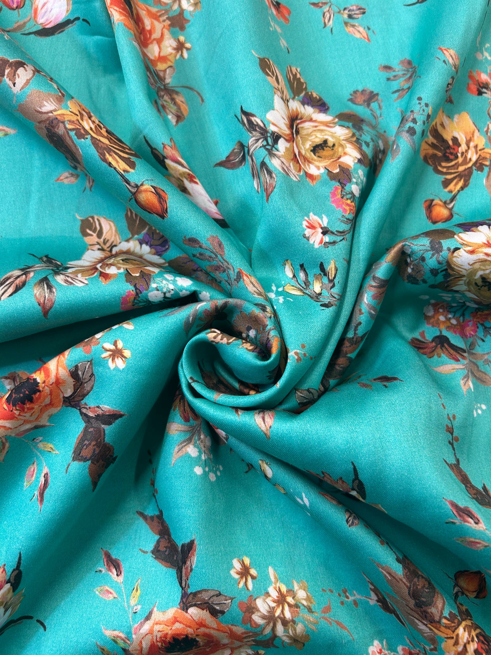 A close-up of a teal fabric with an all-over floral print featuring orange, pink, and brown flowers with green and brown leaves.