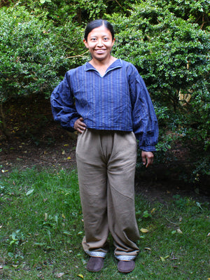 A smiling woman standing outdoors in front of green bushes, wearing a blue long-sleeve collared shirt and loose brown pants with slip-on shoes.