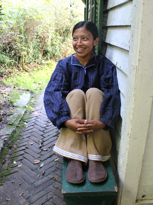 A smiling woman sits on a green wooden bench on a brick path beside a house, wearing a dark blue striped long-sleeve top, tan pants with white lace trim, and brown slippers.