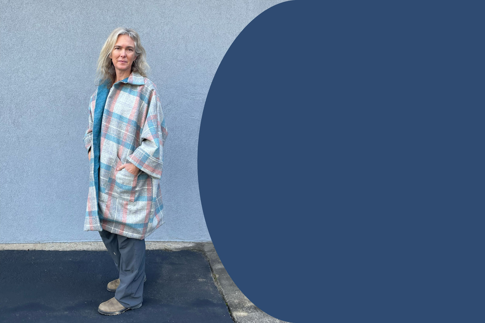 Woman wearing a blue, grey, and pink plaid coat that falls to her knees, outside in front of a blue wall.  Blue block to the right side.