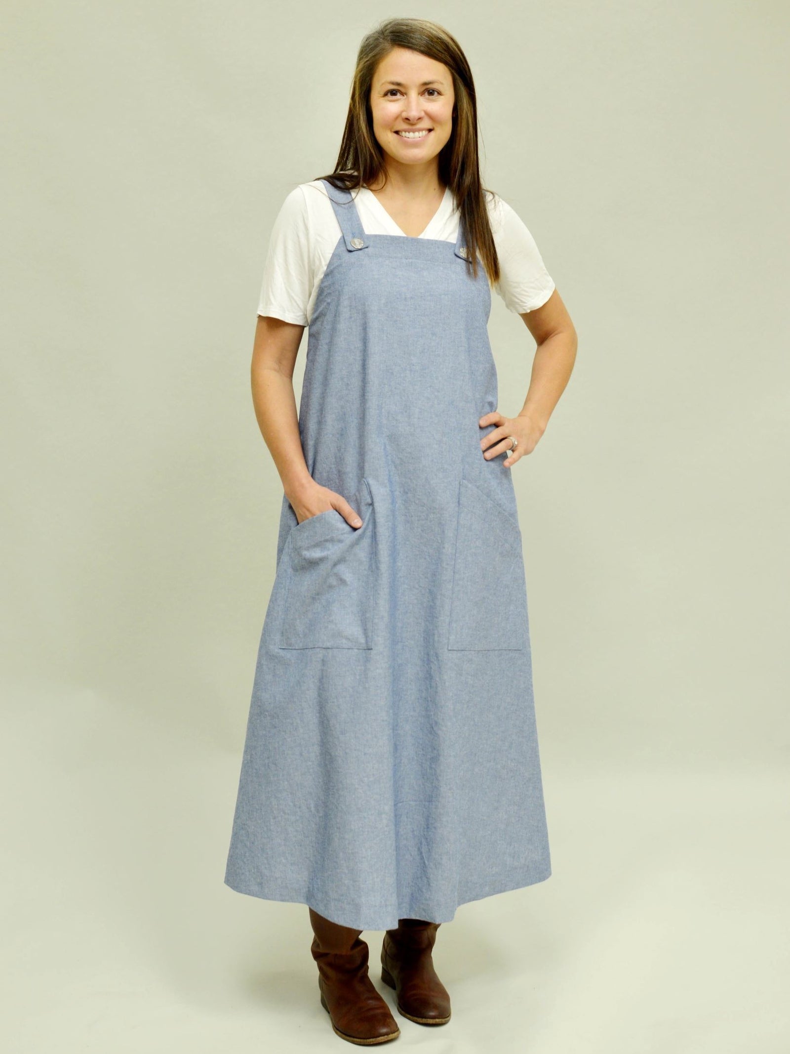 Woman wearing a blue pinafore dress over a white t-shirt in front of a white screen.  Dress has pockets and hand is in one pocket.