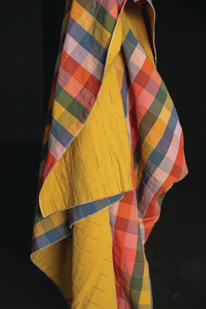 Colorful checked print of yellow, oranges, blues, purples, and pinks  draped over a yellow garment on a black background