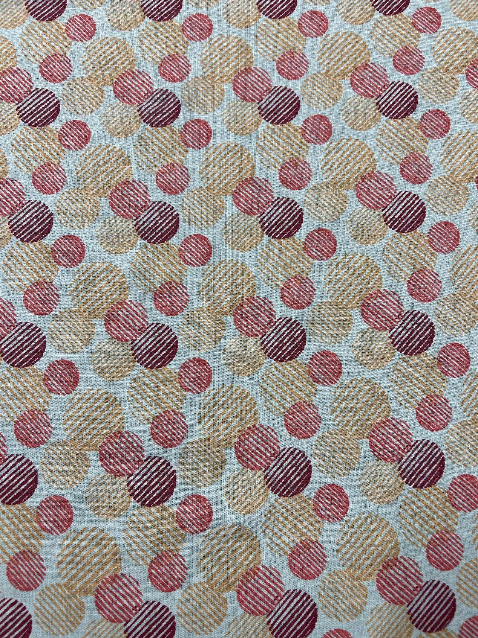  Italian linen with a retro abstract polka dot print fabric in orange, red, and burgundy on an ivory background. The circles vary in size and are filled with fine diagonal stripe textures, creating a layered geometric look with a hand-printed feel. 