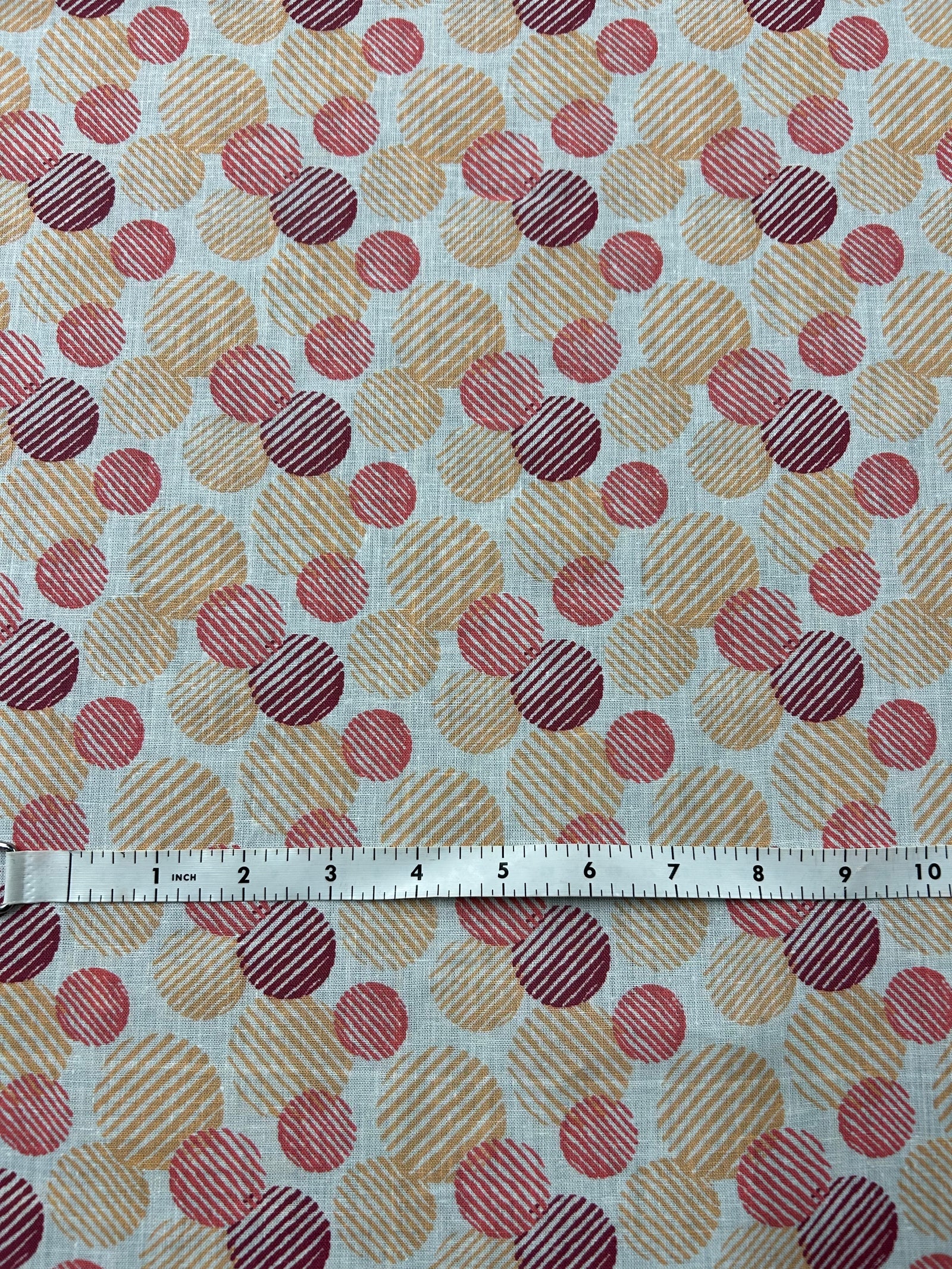  Italian linen with a retro abstract polka dot print fabric in orange, red, and burgundy on an ivory background. The circles vary in size and are filled with fine diagonal stripe textures, creating a layered geometric look with a hand-printed feel. 