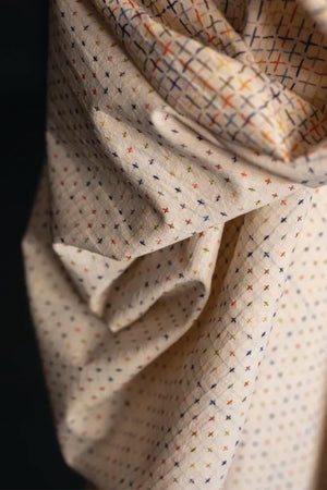 Close up of Indian cotton fabric in ecru base color with cross stitch detailing in multicolor woven greens, blues, orange, yellow and pink.