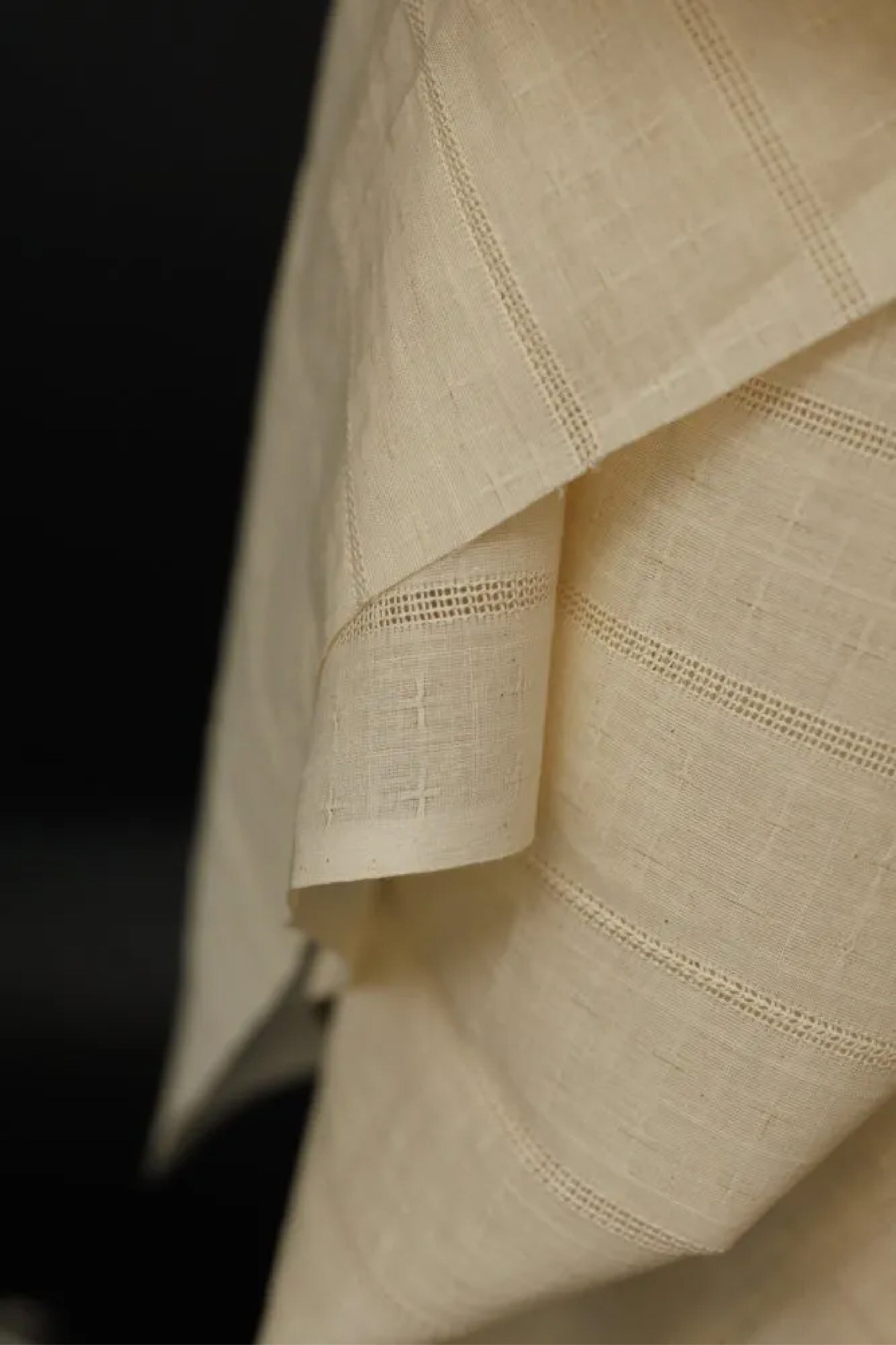 A close up of a draped Indian Cotton beige fabric that is lightweight and semi sheer. The pattern consists of vertical eyelet weaves and woven crosses on a black background.