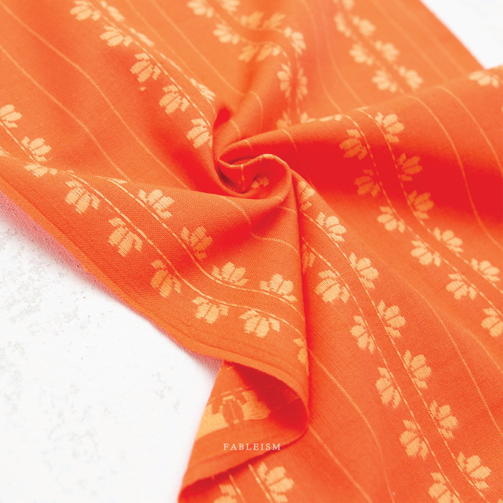 100% cotton fabric in a vibrant stunning orange color with a cream woven textured floral motif arranged in vertical rows in a swirl, on a white background.